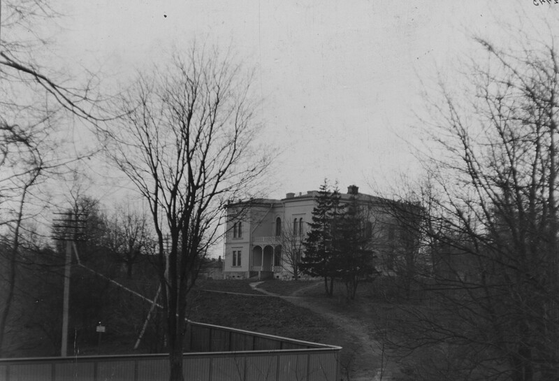 Vaalea kaksikerroksinen rakennus, edessä muutamia puita. Rakennukseen johtaa kapea polku.