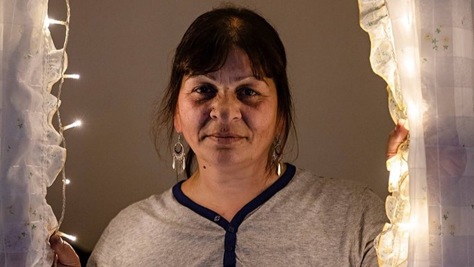 Aikuinen nainen katsoo suoraan kameraan seisten puoliksi avattujen pitsiverhojen välissä, pitäen verhoja käsissään; himmeä lämmin valaistus ja pienet koristevalot luovat kodikkaan tunnelman.