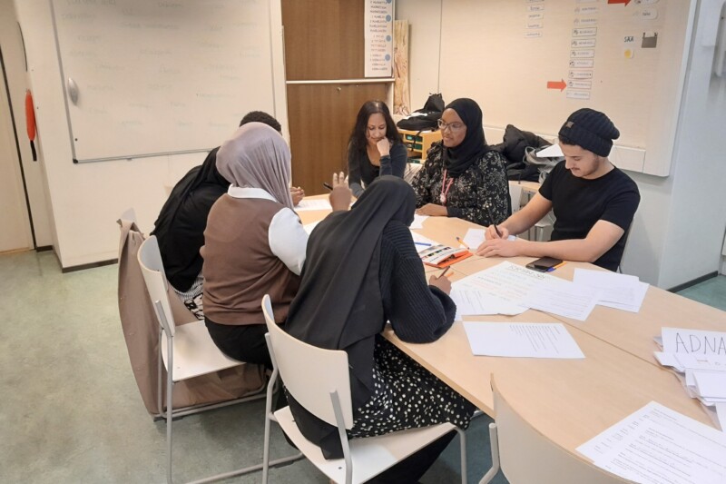 Viisi opiskelijaa istuu luokkahuoneen pöydän ympärillä, kirjoittavat ja keskustelevat papereiden ja muistiinpanojen äärellä; osa käyttää huivia.