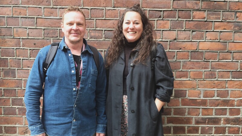 Kaksi henkilöä seisoo tiiliseinän edessä, molemmat hymyilevät ja katsovat kameraan.