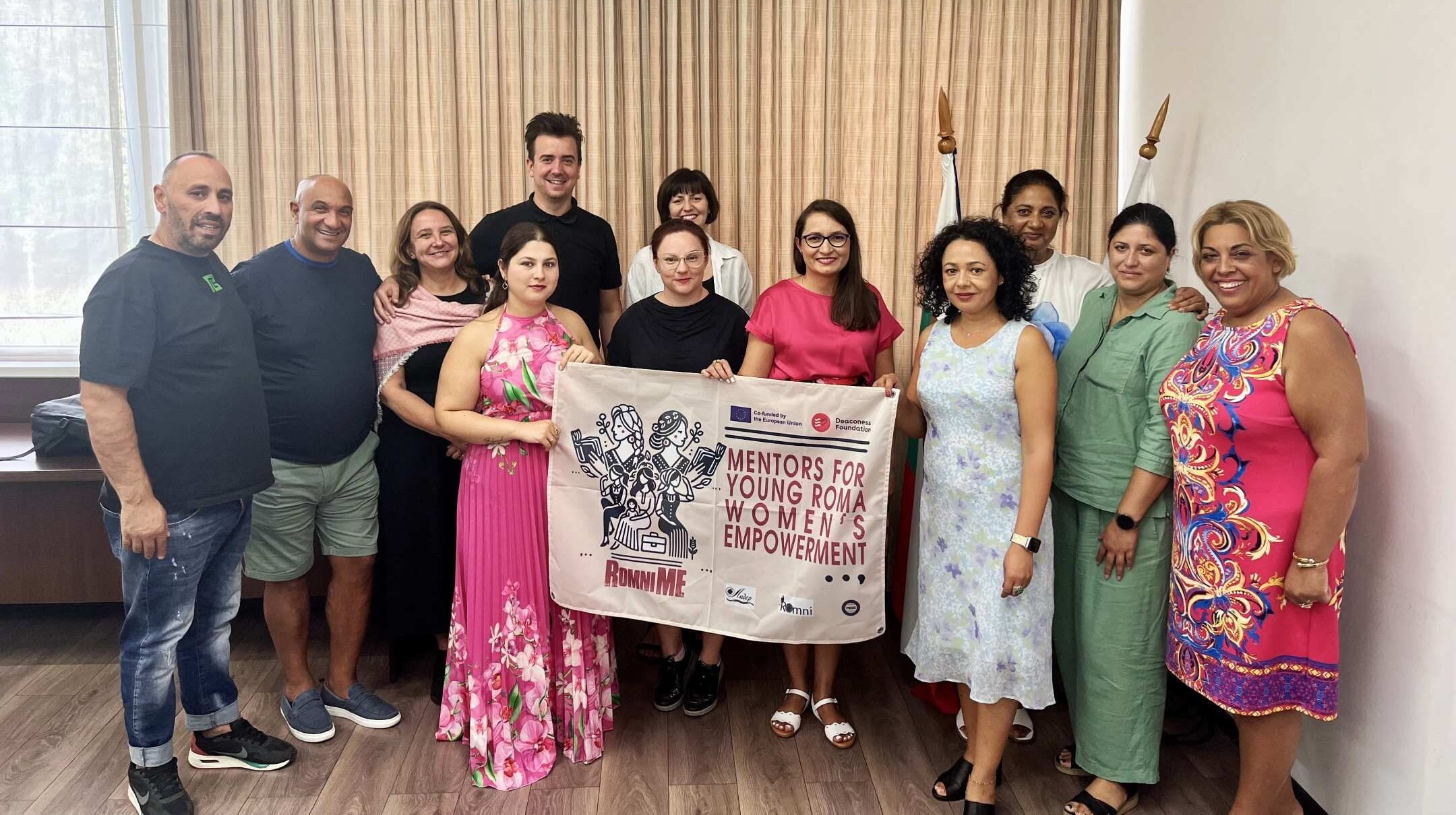 Ryhmä ihmisiä seisoo sisätiloissa ja pitää kädessään kylttiä, jossa lukee "Mentors for Young Roma Women Empowerment".
