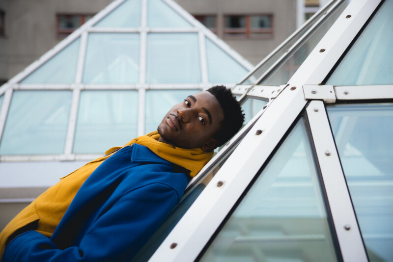 Nuori mies nojaa modernin lasirakenteen kylkeen kaupunkimaisemassa.