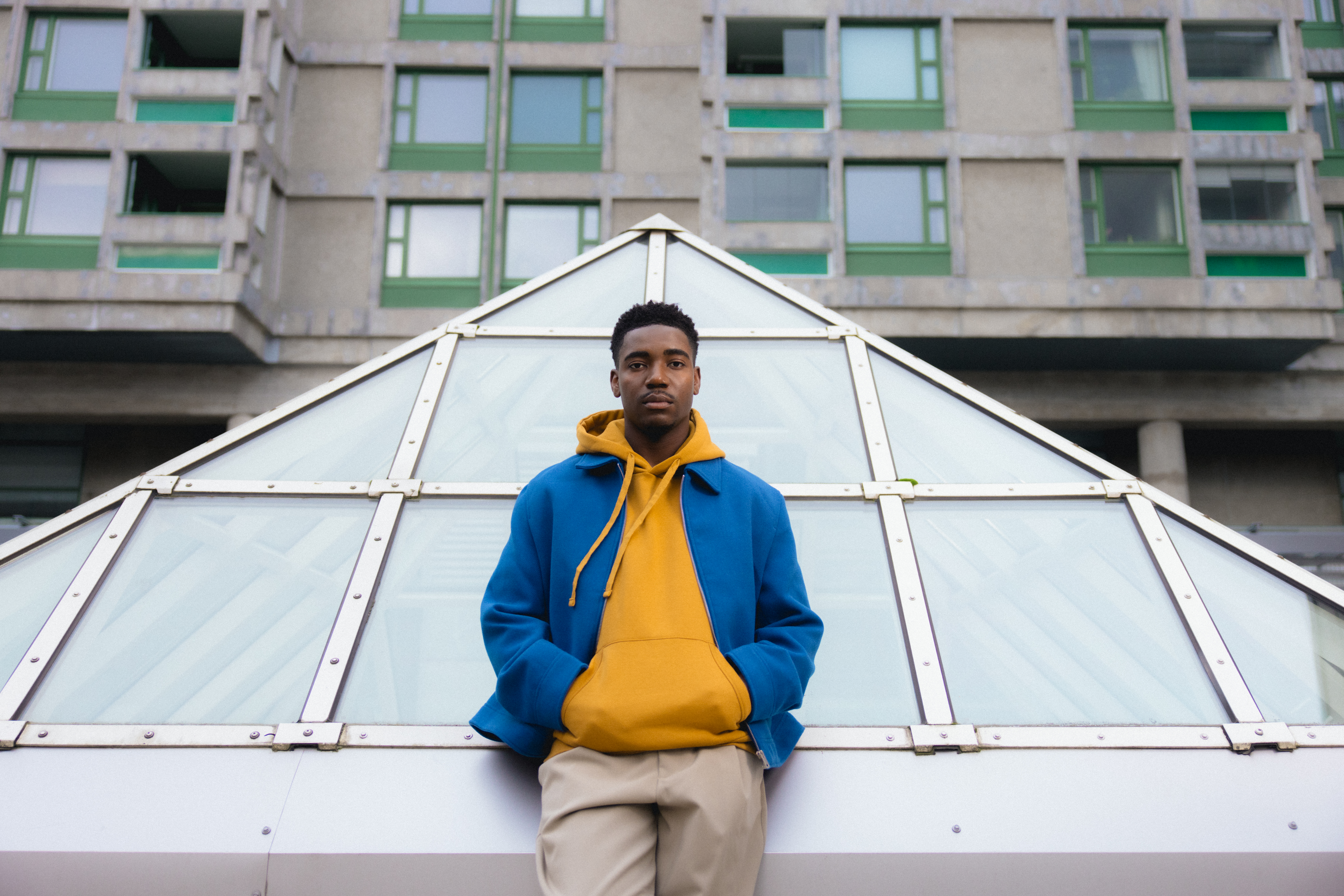 Nuori mies sinisessä takissa ja keltaisessa hupparissa seisoo modernin lasikattoisen rakennuksen edessä urbaanissa ympäristössä.