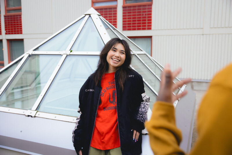 Hymyilevä nuori nainen seisoo ulkona lasikattorakenteen edessä.