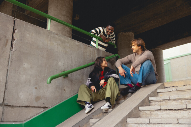 Kolme ihmistä keskustelee rennosti betonirakenteiden ympäröimällä portaikolla, kaksi istuu portailla ja yksi nojaa kaiteeseen yläpuolella.