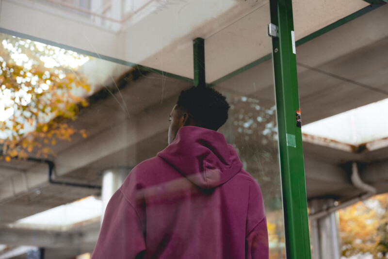 Kuvassa näkyy henkilö takaa päin, jolla on päällään viininpunainen huppari, hän katsoo ulos rakennuksen ikkunasta, taustalla näkyy syksyisiä puita ja betonirakenteita.