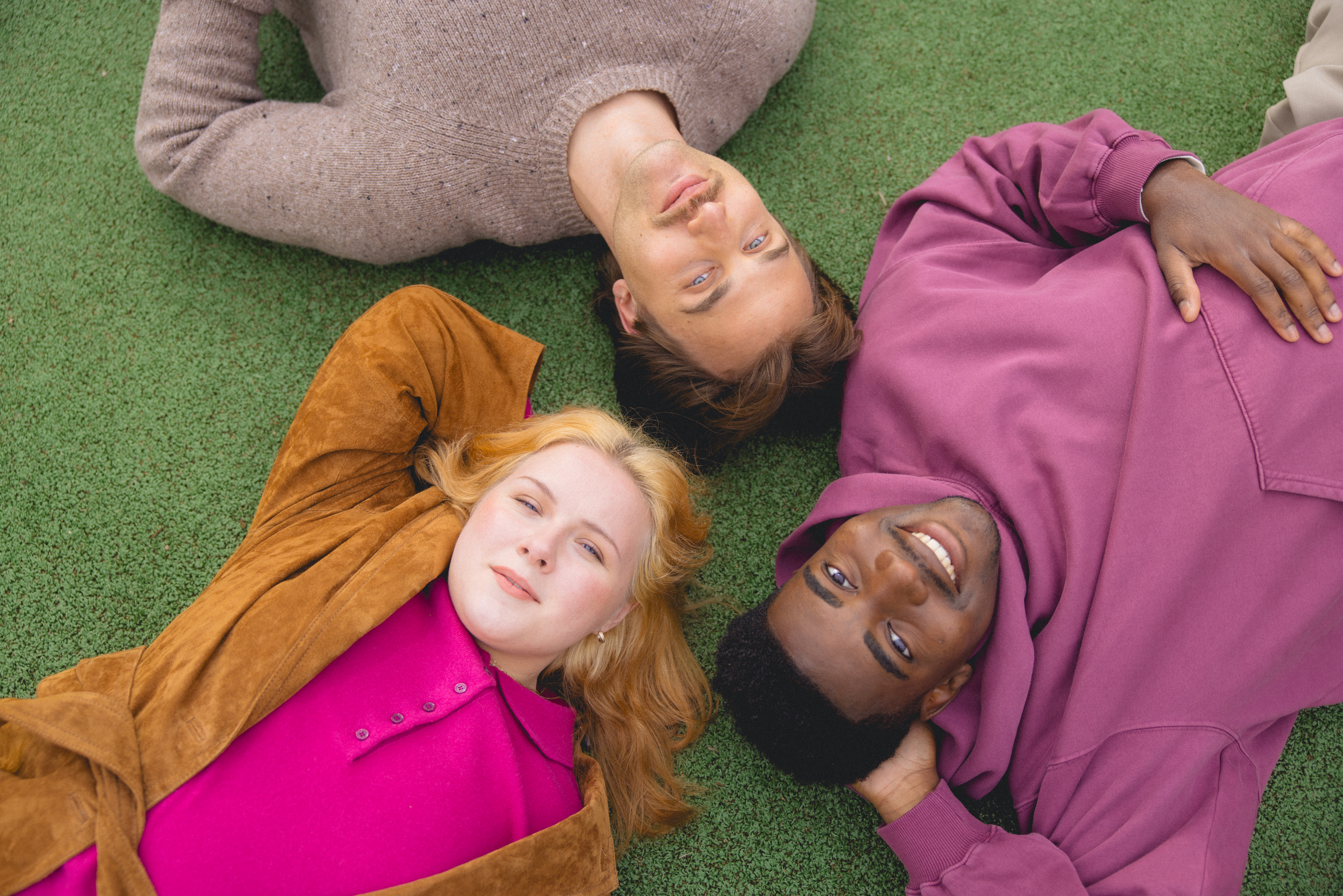Kolme nuorta ihmistä makaa nurmikolla päät yhdessä, katsovat ylöspäin ja hymyilevät.