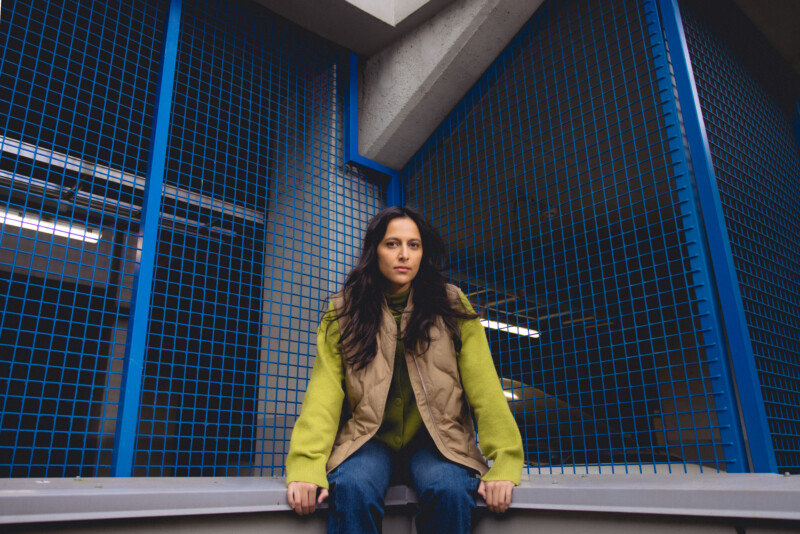 Nuori nainen istuu betonipenkillä sinisten metalliverkkojen edessä, katsoo suoraan kameraan.