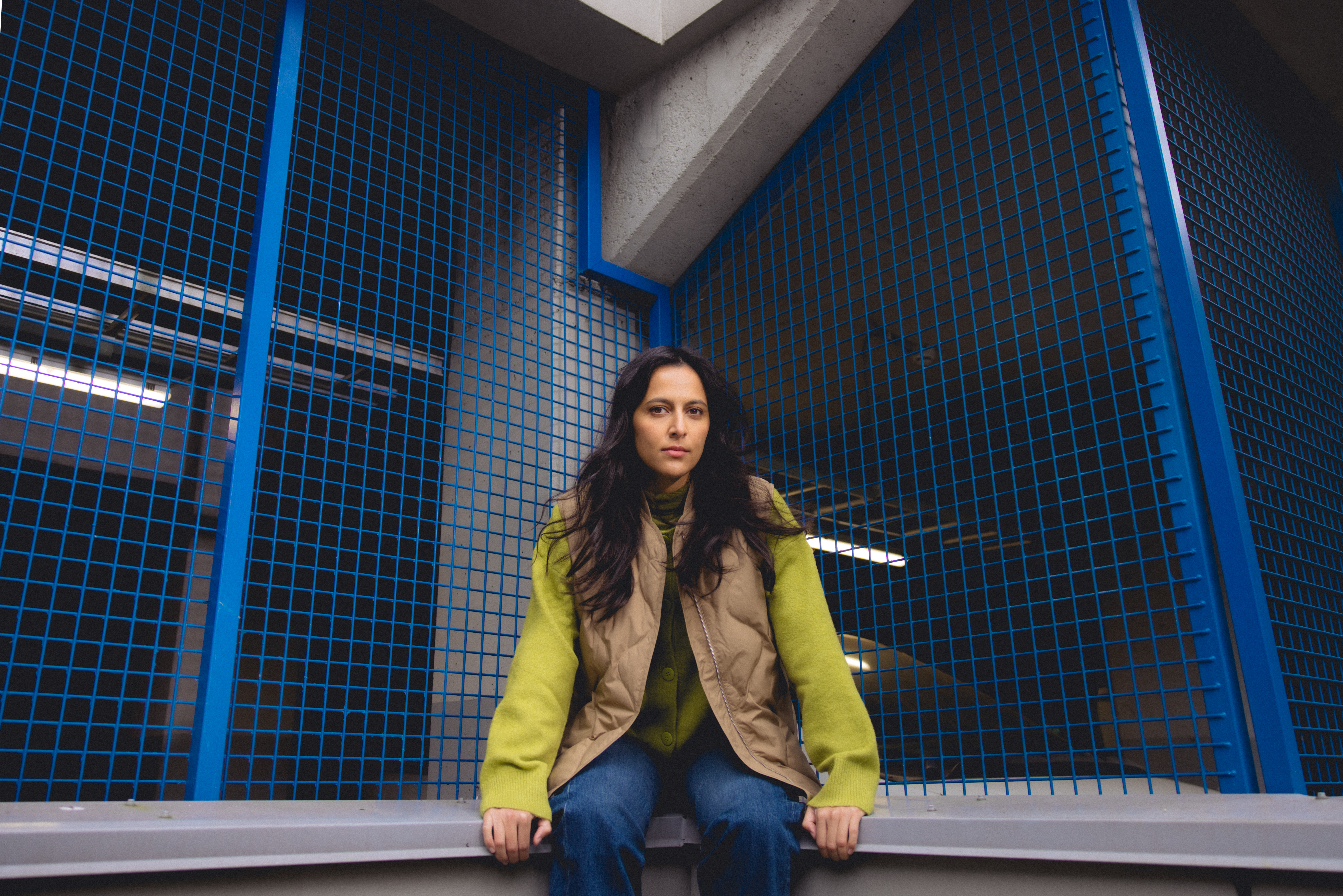 Nuori nainen istuu betonipenkillä sinisten metalliverkkojen edessä, katsoo suoraan kameraan.
