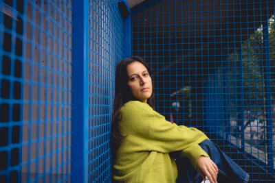 Nuori nainen istuu sinisen metalliverkon edessä ja katsoo kameraan vakavana, hänellä on päällään vihreä neulepusero.