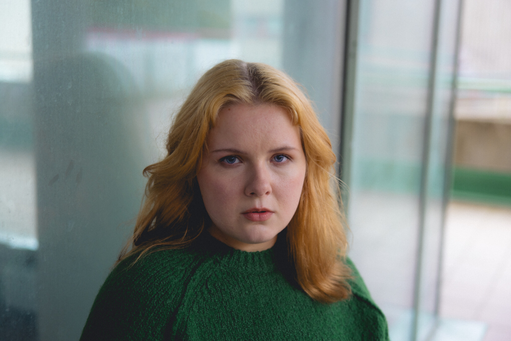 Nuori nainen vihreässä neuleessa katsoo suoraan kameraan, taustalla sumuinen ikkuna.