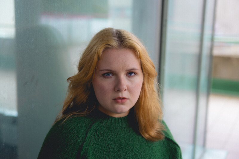 Nuori nainen vihreässä neuleessa katsoo suoraan kameraan, taustalla sumuinen ikkuna.