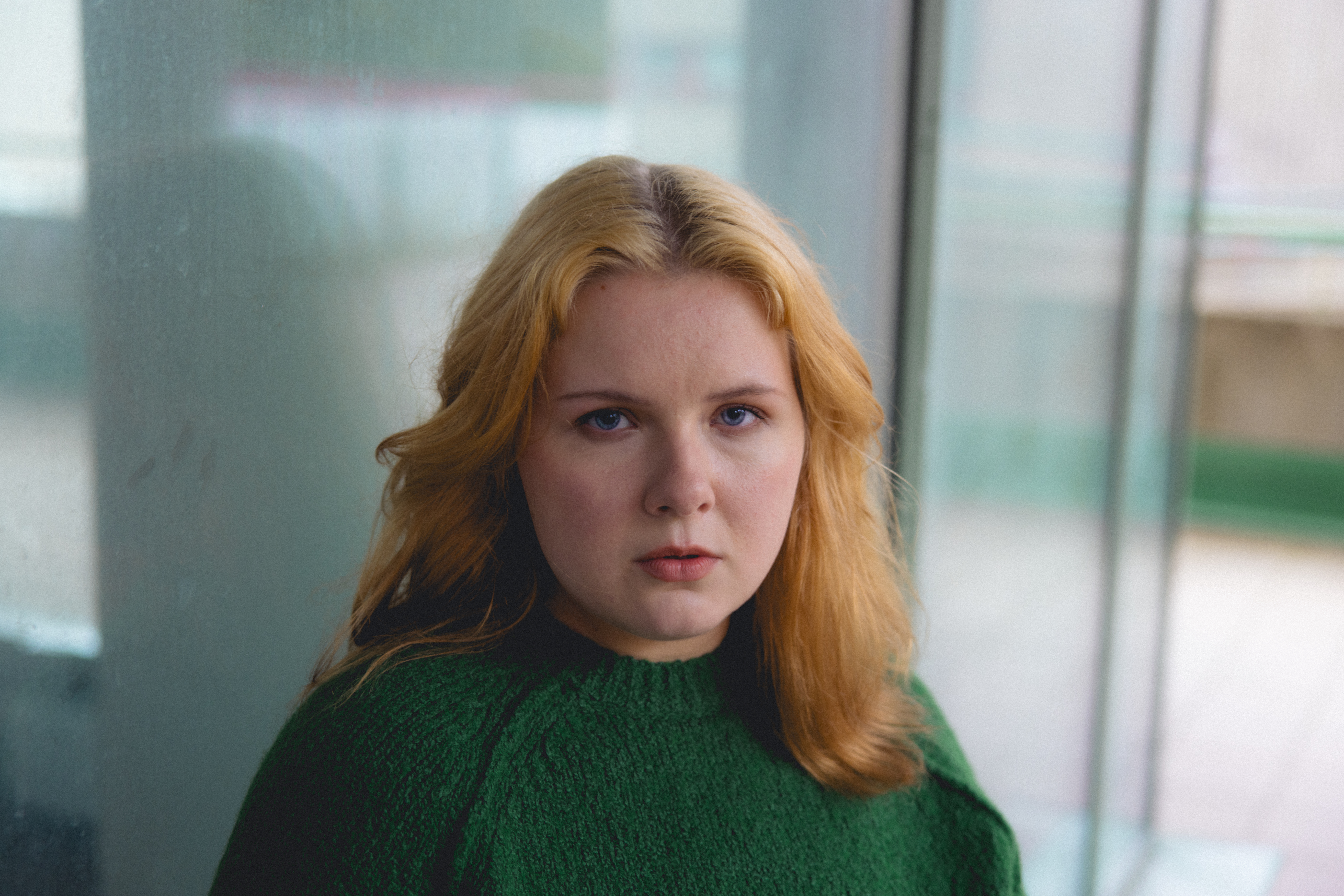 Nuori nainen vihreässä neuleessa katsoo suoraan kameraan, taustalla sumuinen ikkuna.