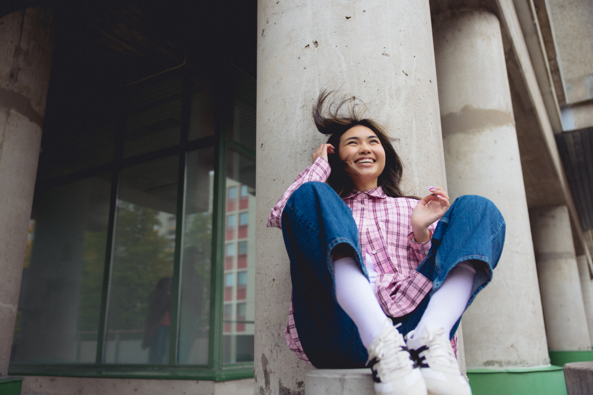 Nuori hymyilevä nainen istuu pylvään vieressä ulkona, pukeutuneena ruudulliseen paitaan, sinisiin farkkuihin ja valkoisiin tennareihin.