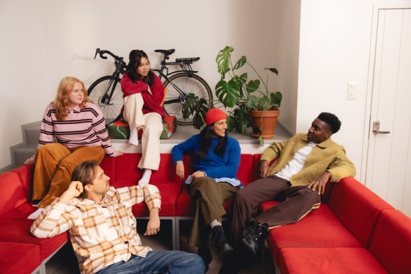 Ryhmän nuoria aikuisia istuu ja keskustelee punaisella sohvallaan sisätiloissa, taustalla näkyy polkupyörä ja viherkasvi.