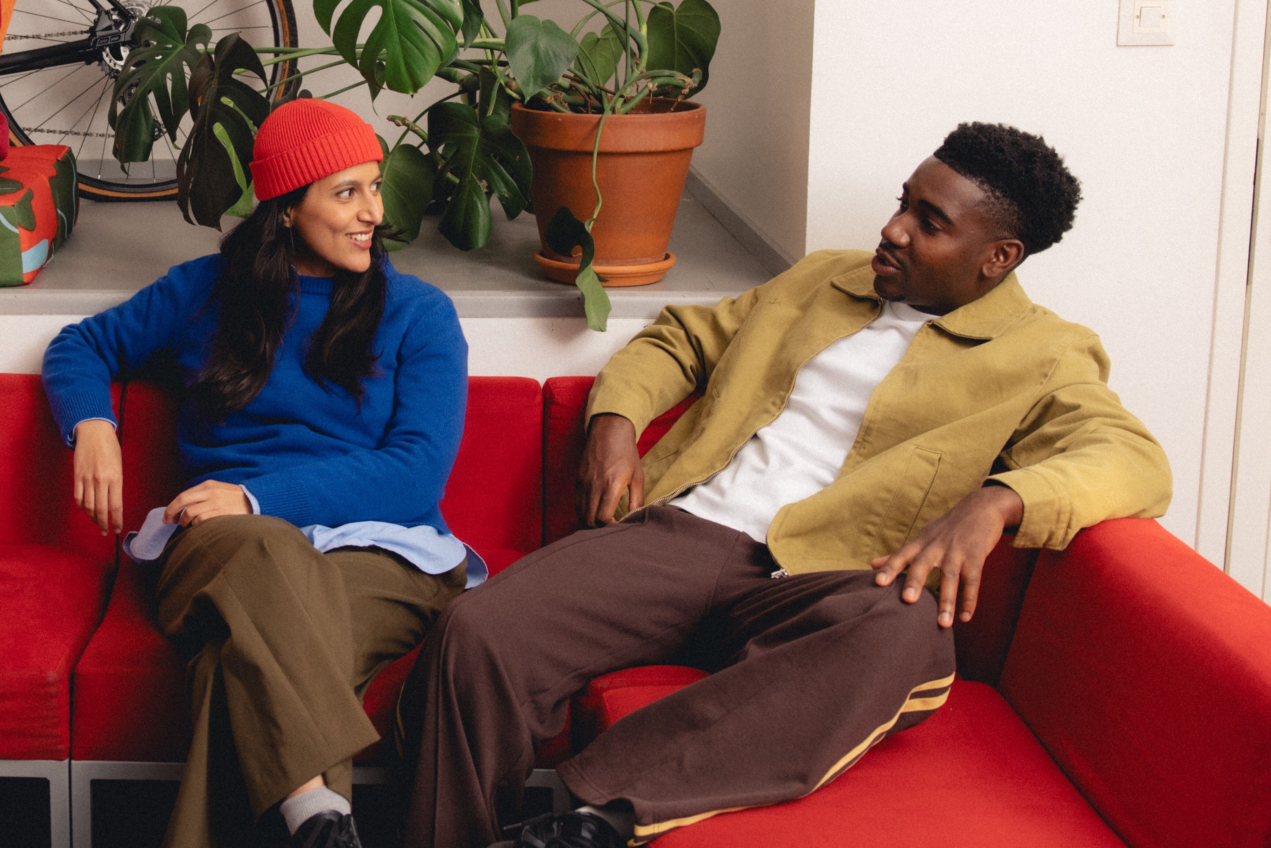 Kaksi nuorta istuu punaisella sohvallla ja keskustelee rennosti sisätiloissa, taustalla viherkasveja ja polkupyörä.
