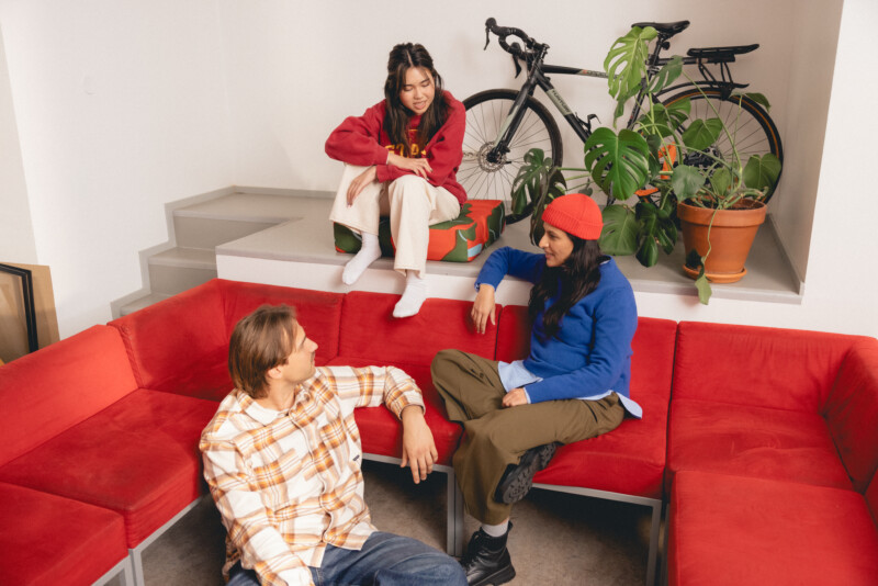 Kolme nuorta istuu ja juttelee punaisella kulmasohvalla ja sen läheisyydessä sisätiloissa, taustalla näkyy polkupyörä ja viherkasveja.