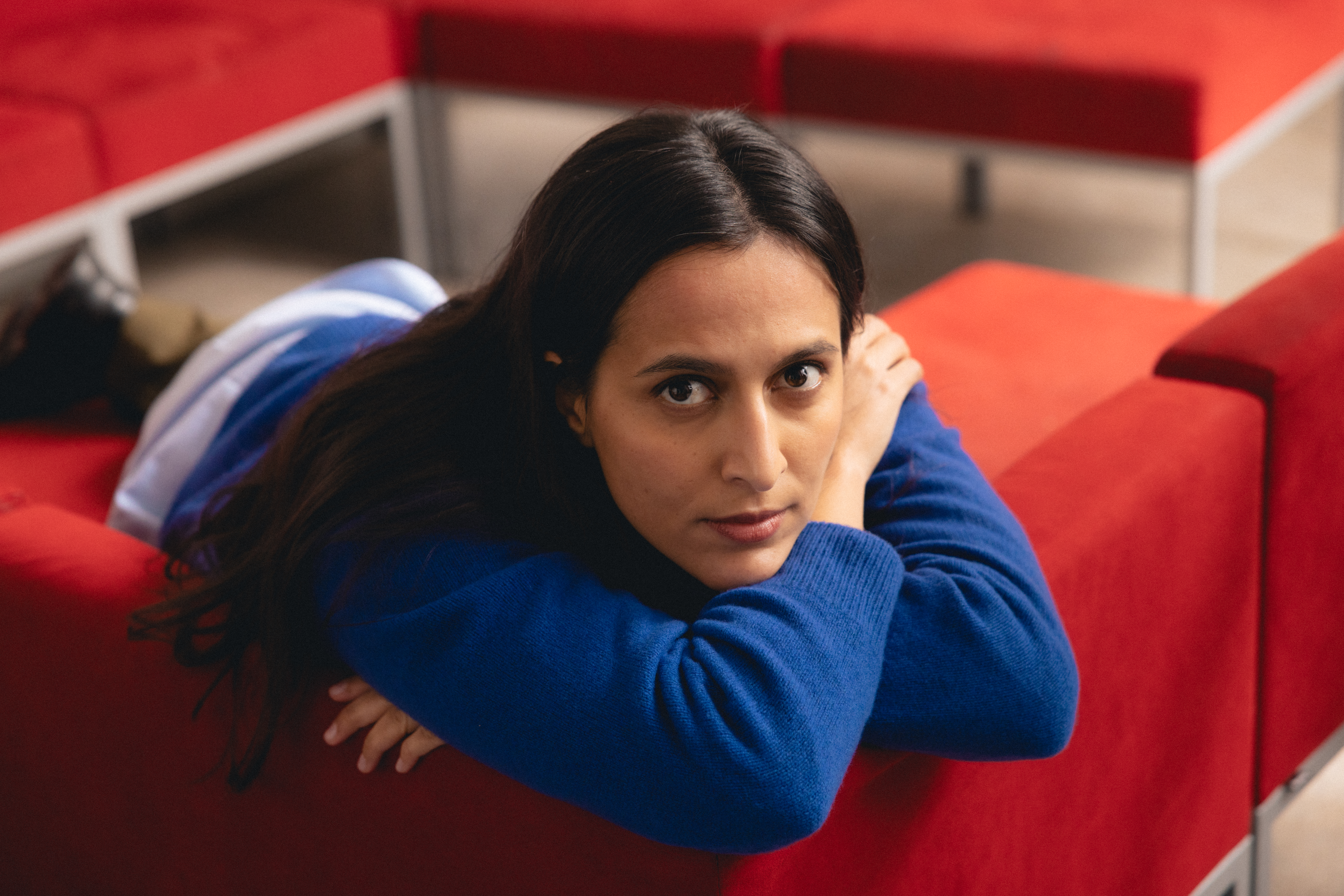 Nainen nojaa käsillään punaisen sohvan selkänojaan ja katsoo suoraan kameraan.