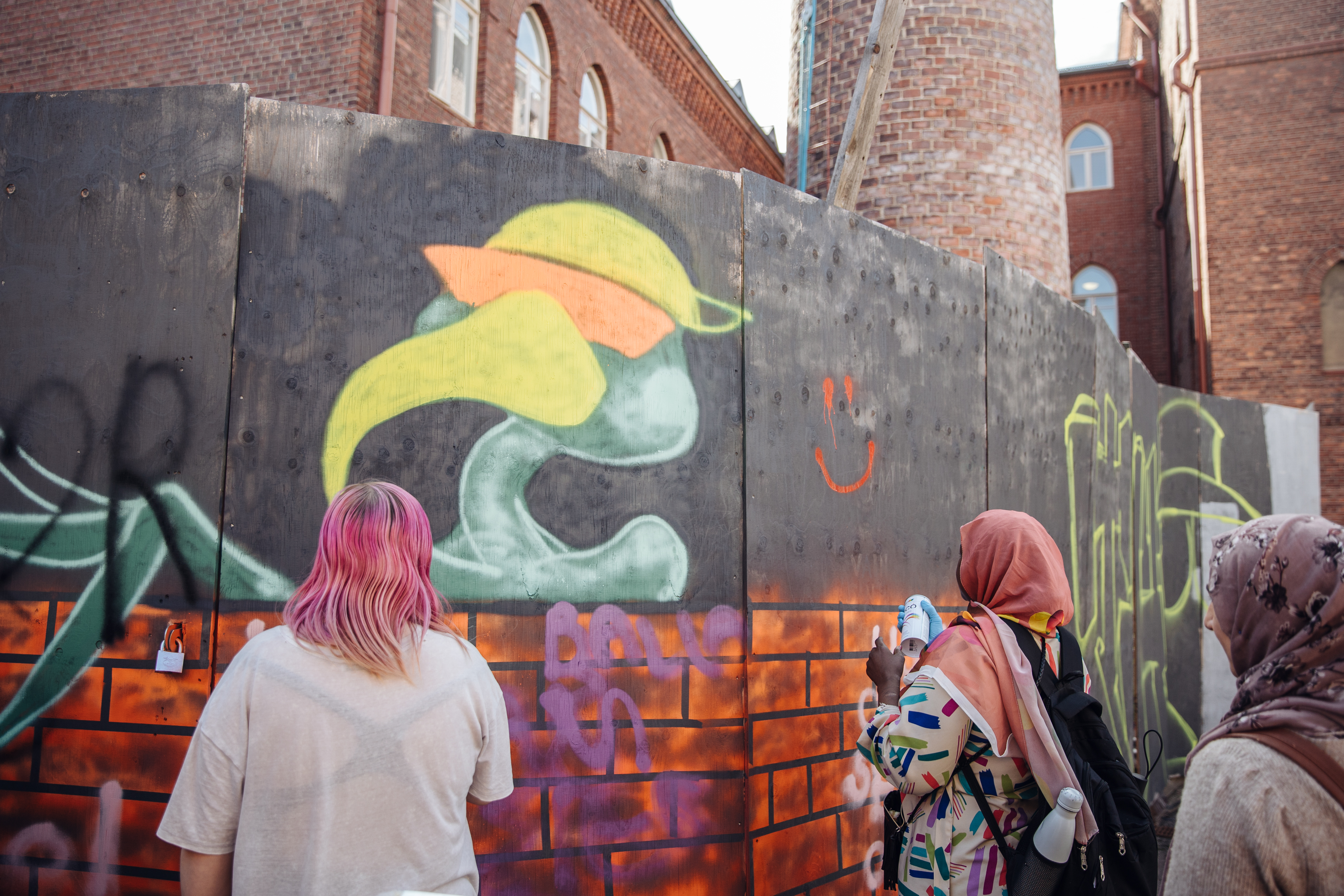 Three people with colorful hair and headscarves spray painting graffiti on a black wall outdoors near a red brick building.