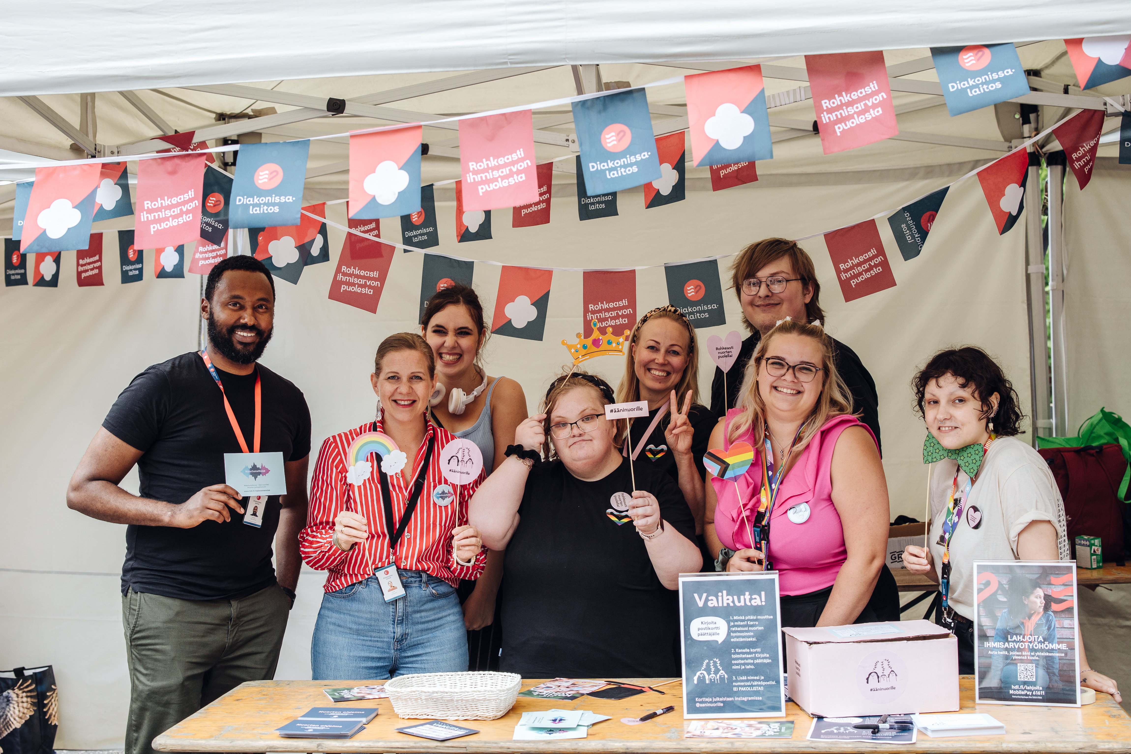 Monikulttuurinen ryhmä hymyileviä nuoria ja aikuisia seisoo teltan alla esittelemässä Ääni nuorille -toimintamallia, ympärillä värikkäitä julisteita ja lippuja.