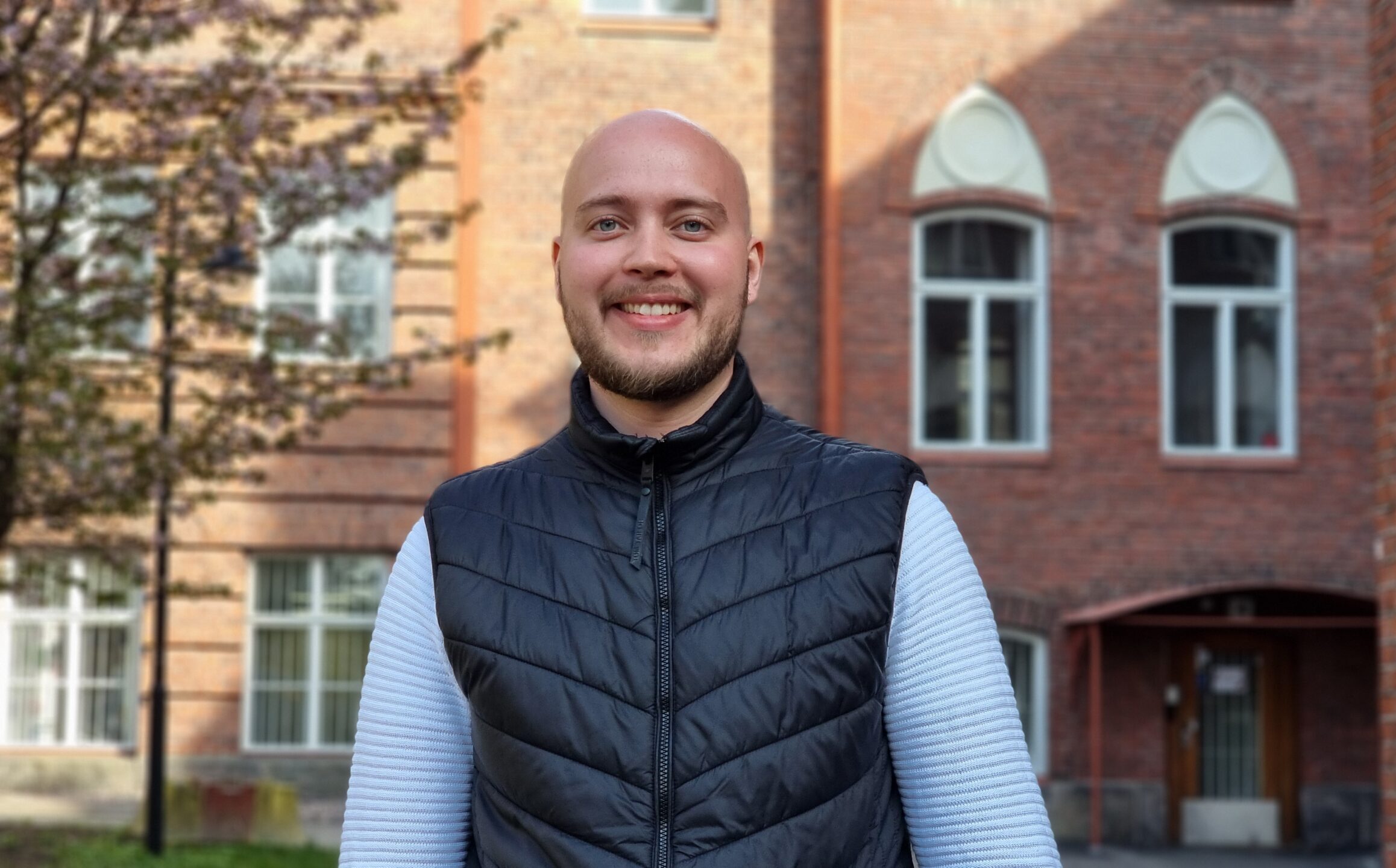 Mies seisoo ulkona, taustalla tiilinen rakennus ja ikkunat, hän hymyilee ja pukeutunut mustaan liiviin ja vaaleaan pitkähihainen puseroon.