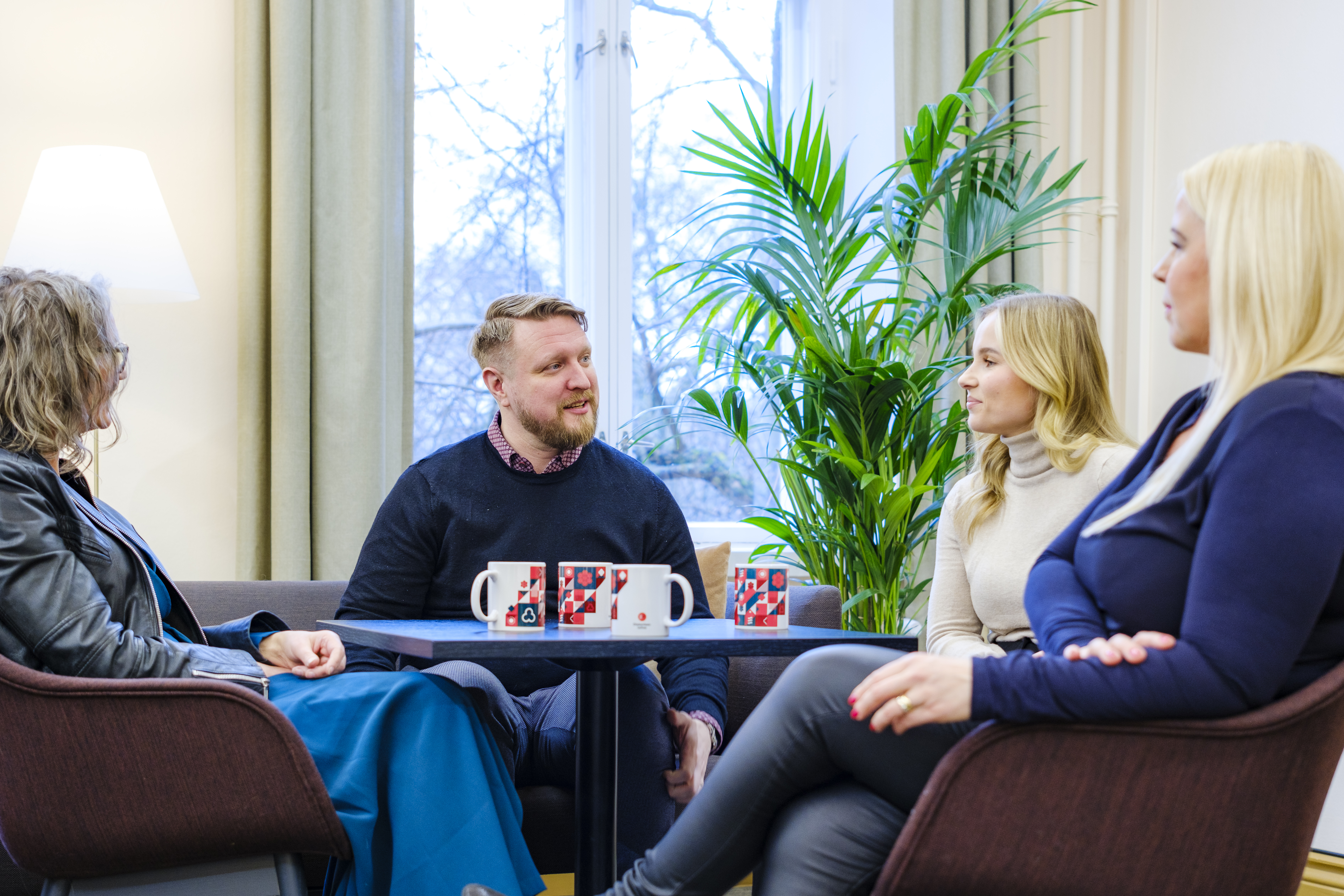 Neljä ihmistä istuu pöydän ympärillä keskustellen kodikkaassa huoneessa, taustalla iso ikkuna ja viherkasvi.