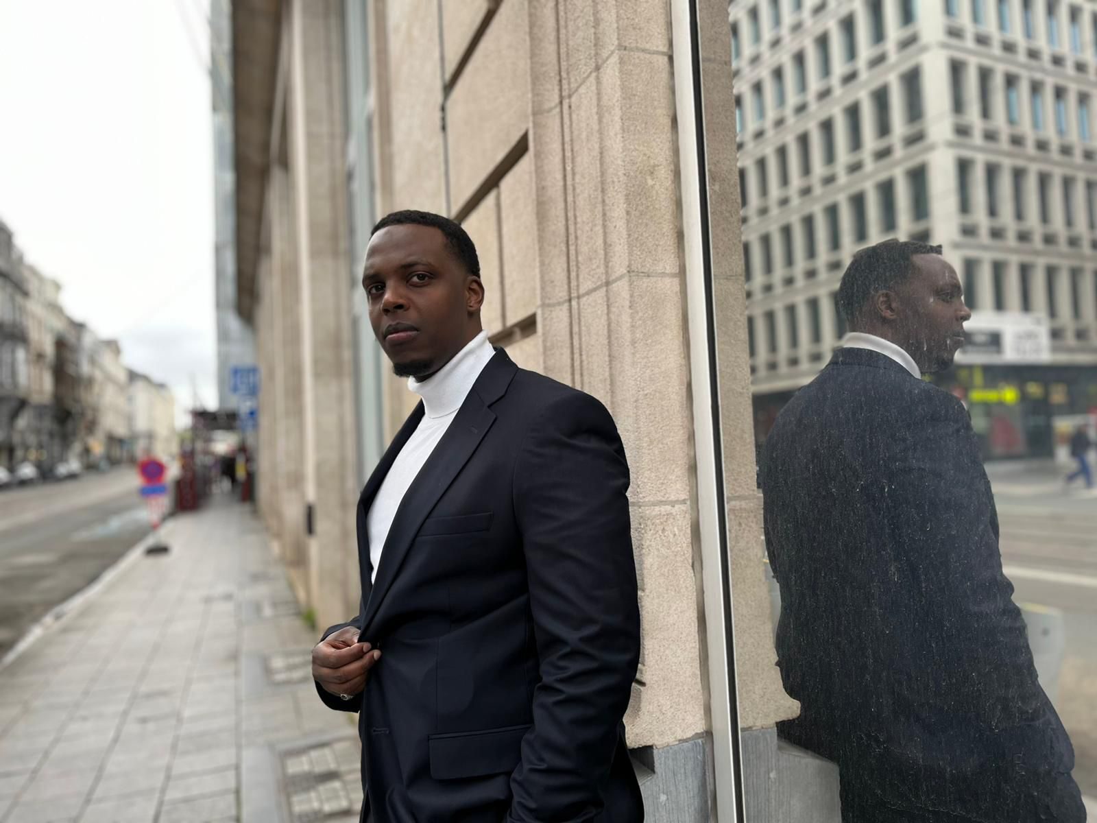 Mies tummahihaisessa puvussa nojaa rakennuksen kulmaan, hänen peilikuvansa näkyy ikkunassa.