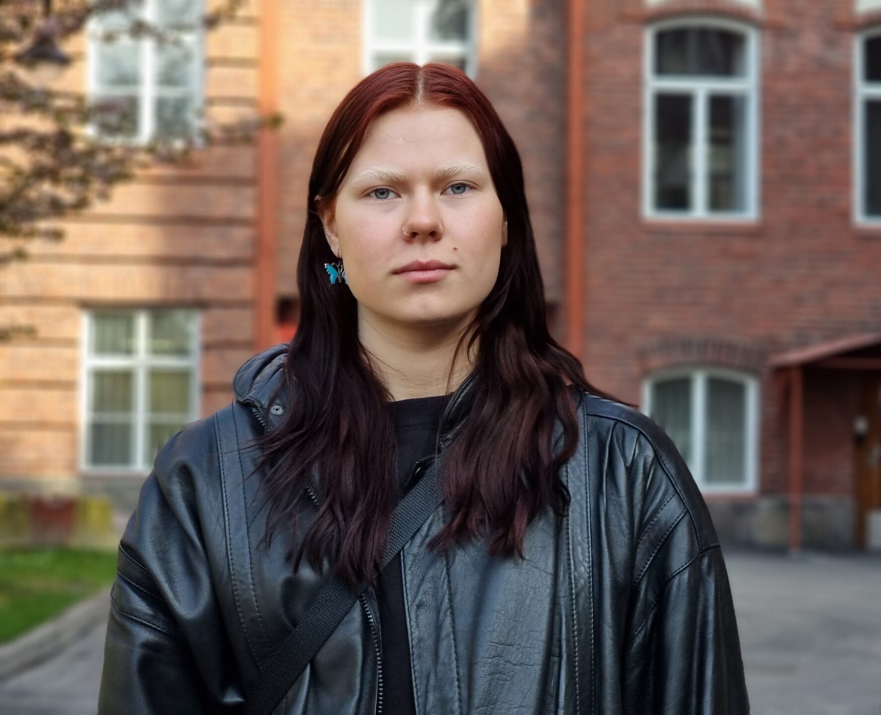 Nuori nainen mustassa nahkatakissa seisoo ulkona rakennuksen edustalla, taustalla näkyy tiiliverhoiltuja ikkunoita.