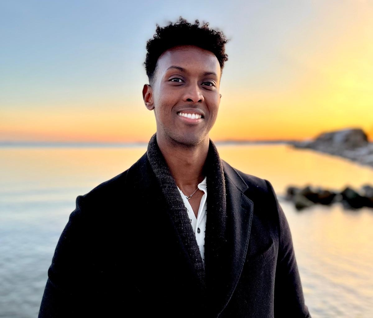 Nuori mies hymyilee ulkona meren rannalla auringonlaskun aikaan, pukeutuneena mustaan takkiin.