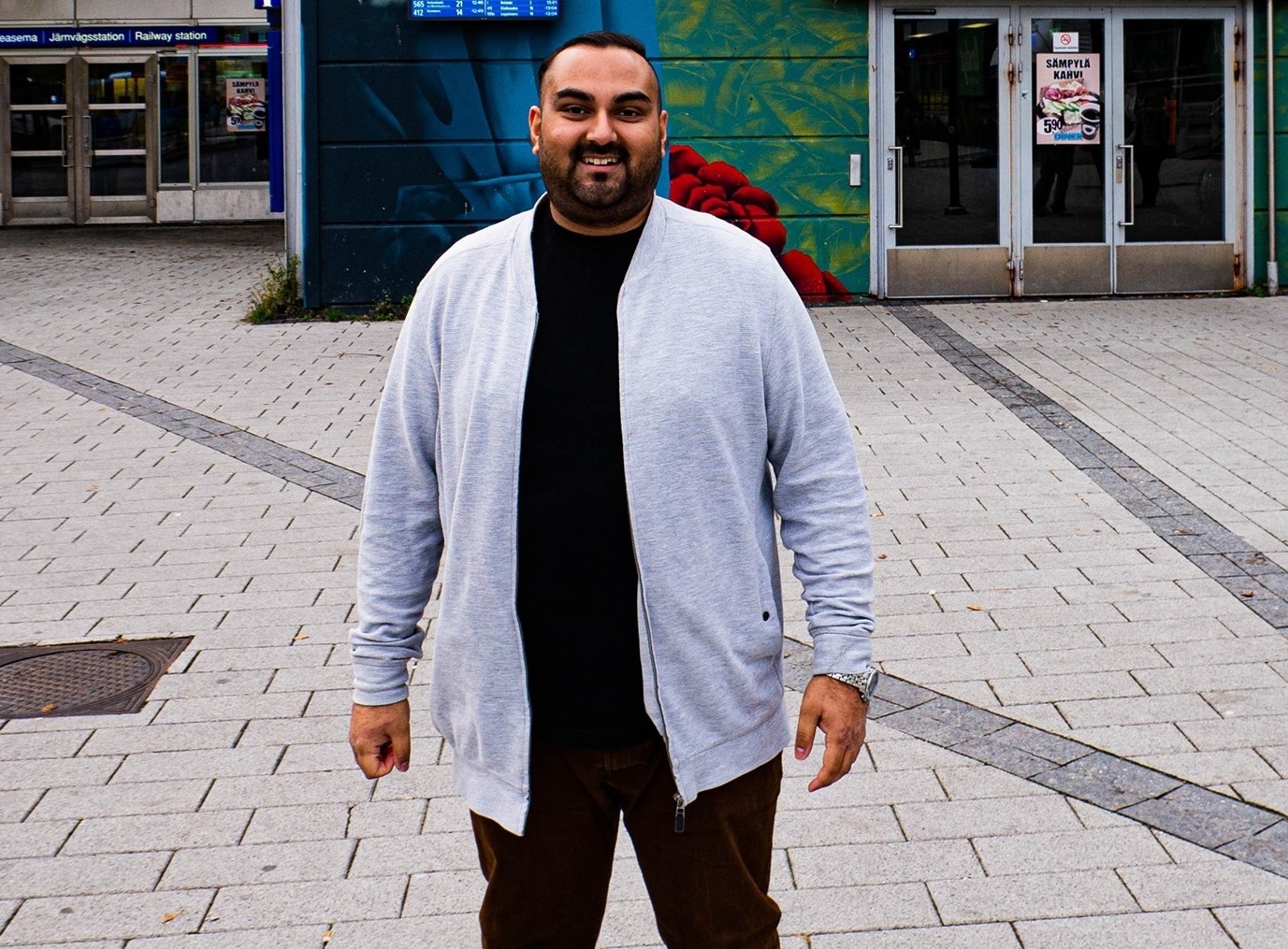 Mies seisoo ulkona kivetyllä aukiolla, hänellä on harmaa neuletakki ja musta paita, taustalla rakennuksen sisäänkäynti ja värikäs seinämaalaus.