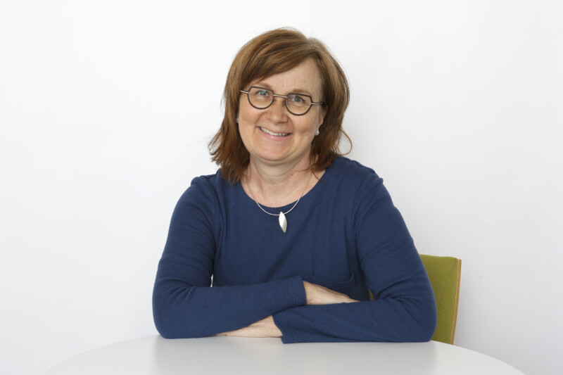 Ruskeahiuksinen, siniseen paitaan pukeutunut nainen katsoo hymyillen kameraan.