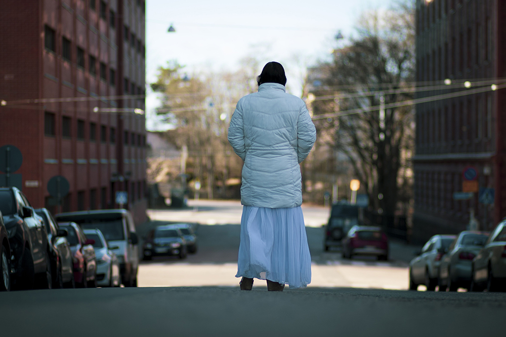 Henkilö seisoo selin kameralle kaupungin kadulla talvitakissa ja pitkässä hameessa, ympärillä kerrostaloja ja pysäköityjä autoja.
