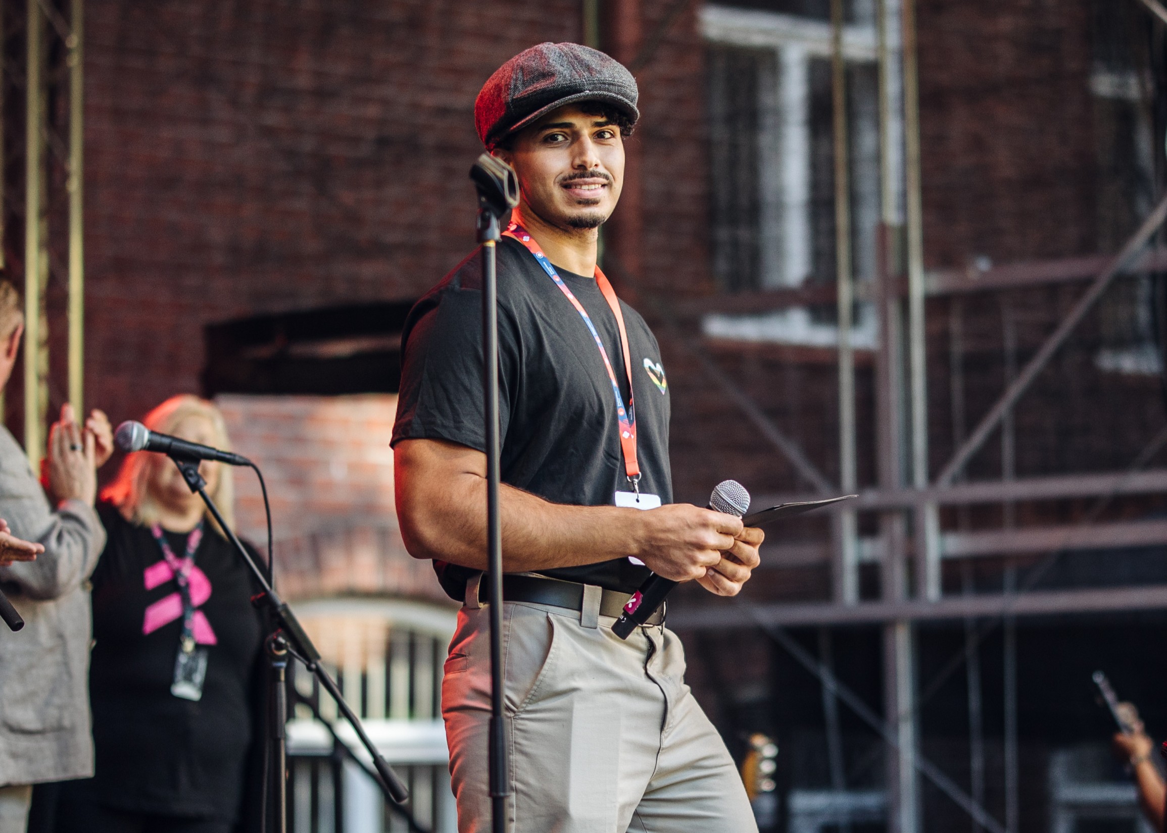 Nuori mies seisoo mikrofonin kanssa ulkoilmalavalla, taustalla ihmisiä ja tiiliseinä.