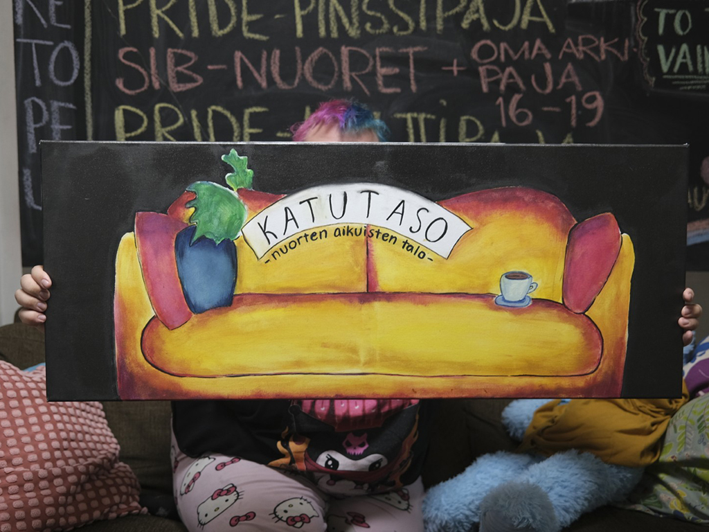 Henkilö istuu värikkäällä taustalla ja pitää käsissään maalattua kylttiä, jossa on keltainen sohva, viinirypäleitä ja kahvikuppi sekä teksti "Katutaso nuorten aikuisten talo".