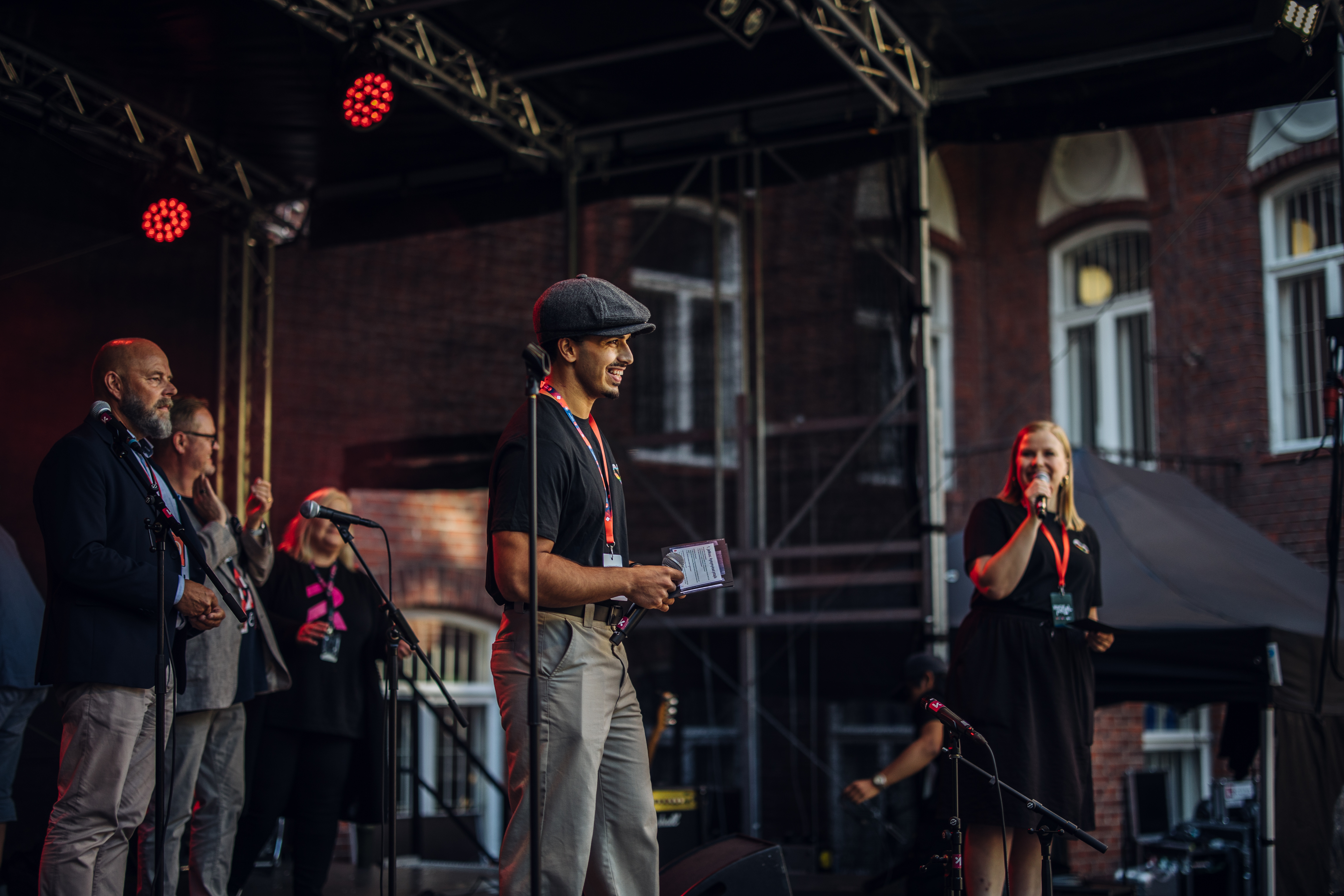 Ihmisiä esiintymislavalla iltavalaistuksessa, yksi mies ja yksi nainen keskellä puhumassa mikrofoneihin, taustalla muita ihmisiä ja rakennuksia.