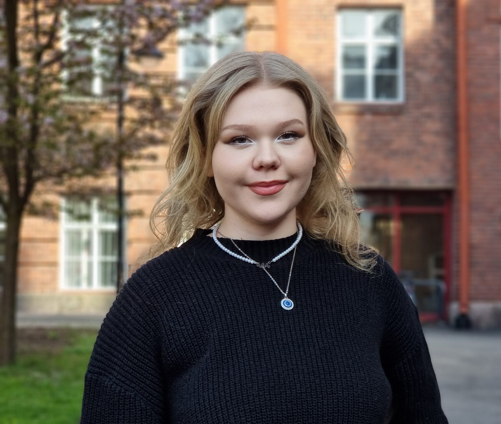 Nuori nainen, joka hymyilee ja katsoo suoraan kameraan, taustalla kerrostalo ja puu.