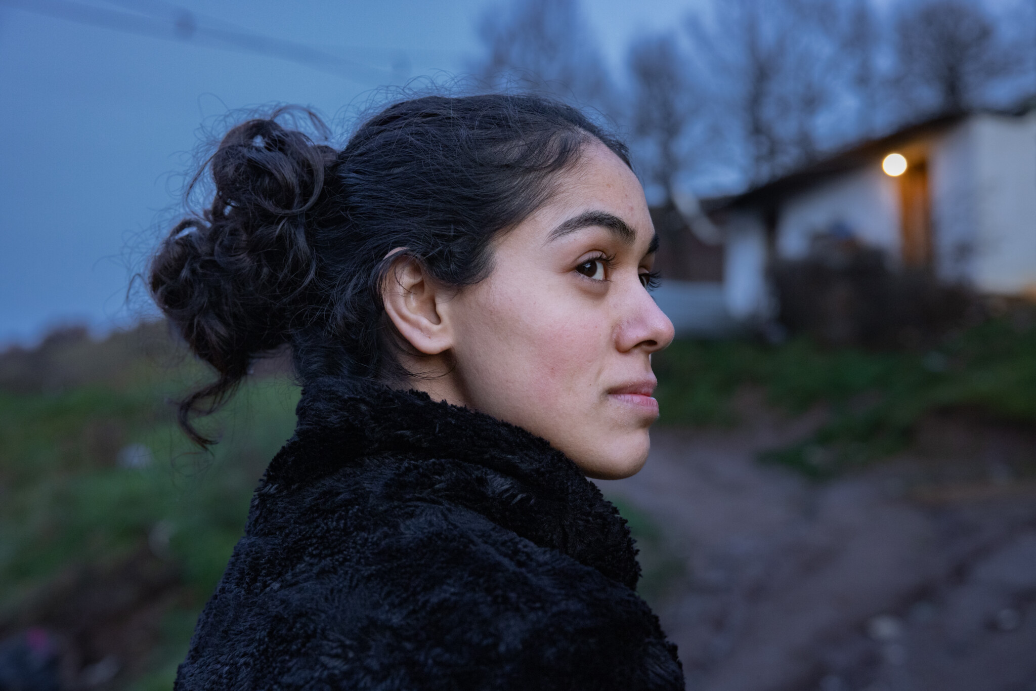 Nuori nainen katsoo sivulle ulkona hämärässä, taustalla epätarkka talo ja luonto.