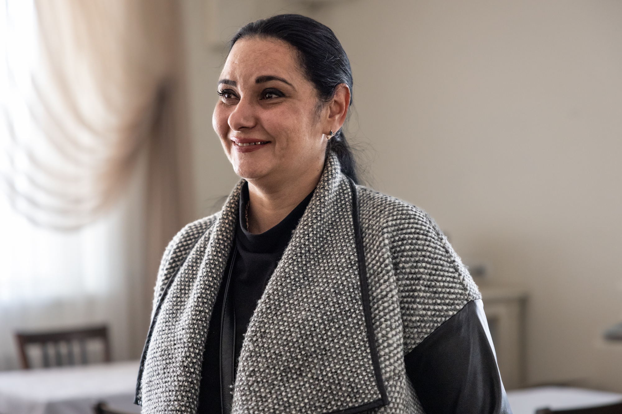 Hymyilevä nainen harmaassa neuletakissa vaaleassa huoneessa.