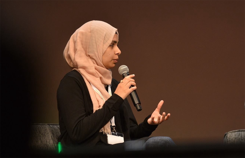 Nainen, jolla vaalea huivi ja musta paita, puhuu mikrofonin kanssa istuen.