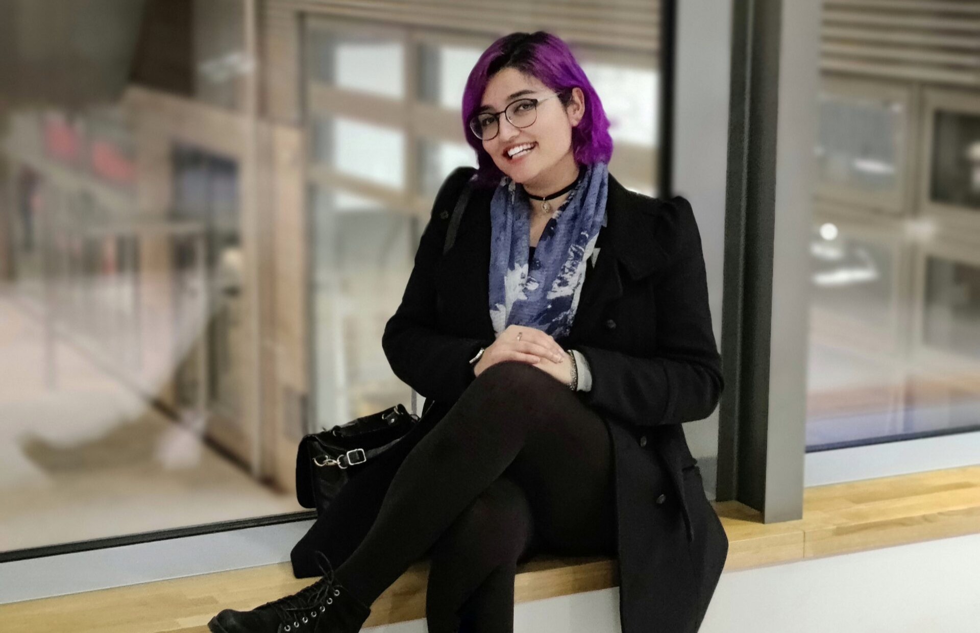 Nuorehko henkilö violetit hiukset, silmälasit ja musta takki istuu hymyillen sisätiloissa ikkunan vieressä.