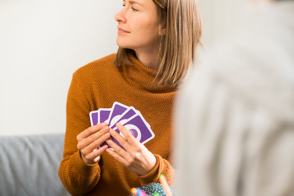 Nainen oranssassa neuleessa istuu ja pitää kädessään kortteja, katsoen sivulle, taustalla osittain näkyvä toinen henkilö.