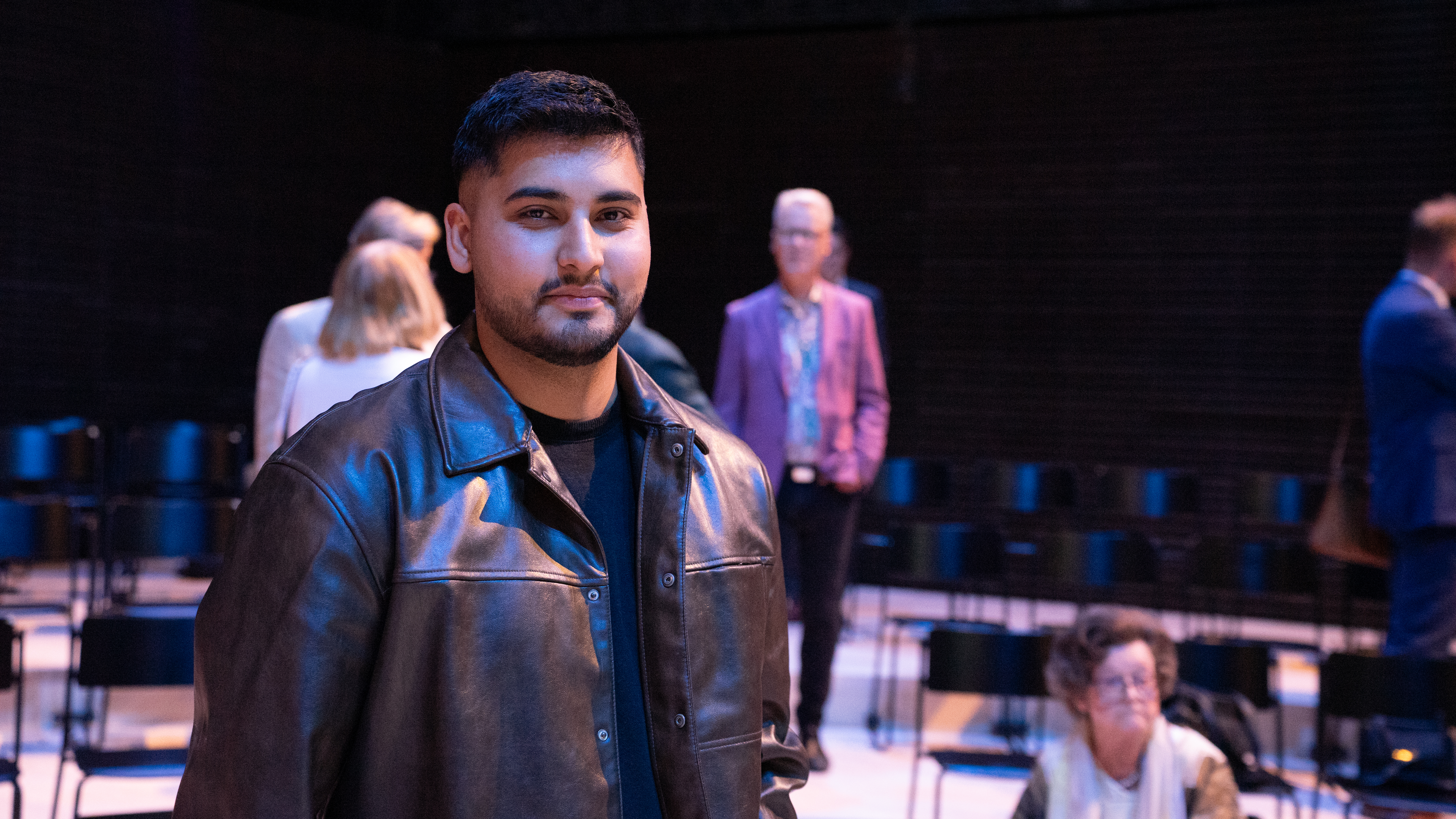 Nuori mies nahkatakissa seisoo hämärässä näyttämöympäristössä, taustalla useita ihmisiä ja tyhjiä tuoleja.