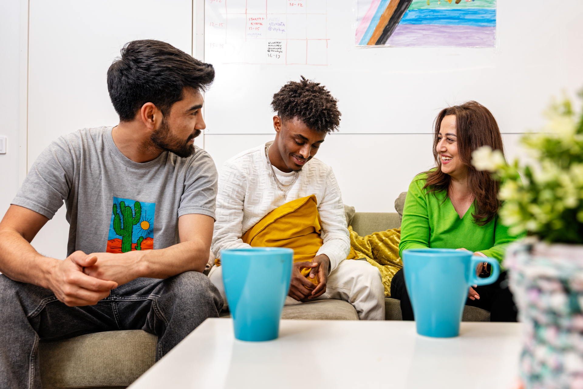 Nuori mies maalaamassa värikästä esinettä pöydän ääressä, taustalla toinen nuori nainen maalaamassa.