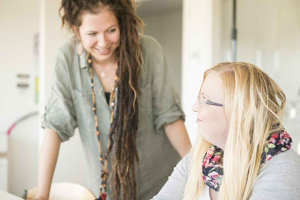 Kaksi naista keskustelee iloisesti sisätiloissa, toinen istuu ja toinen nojaa pöytään.