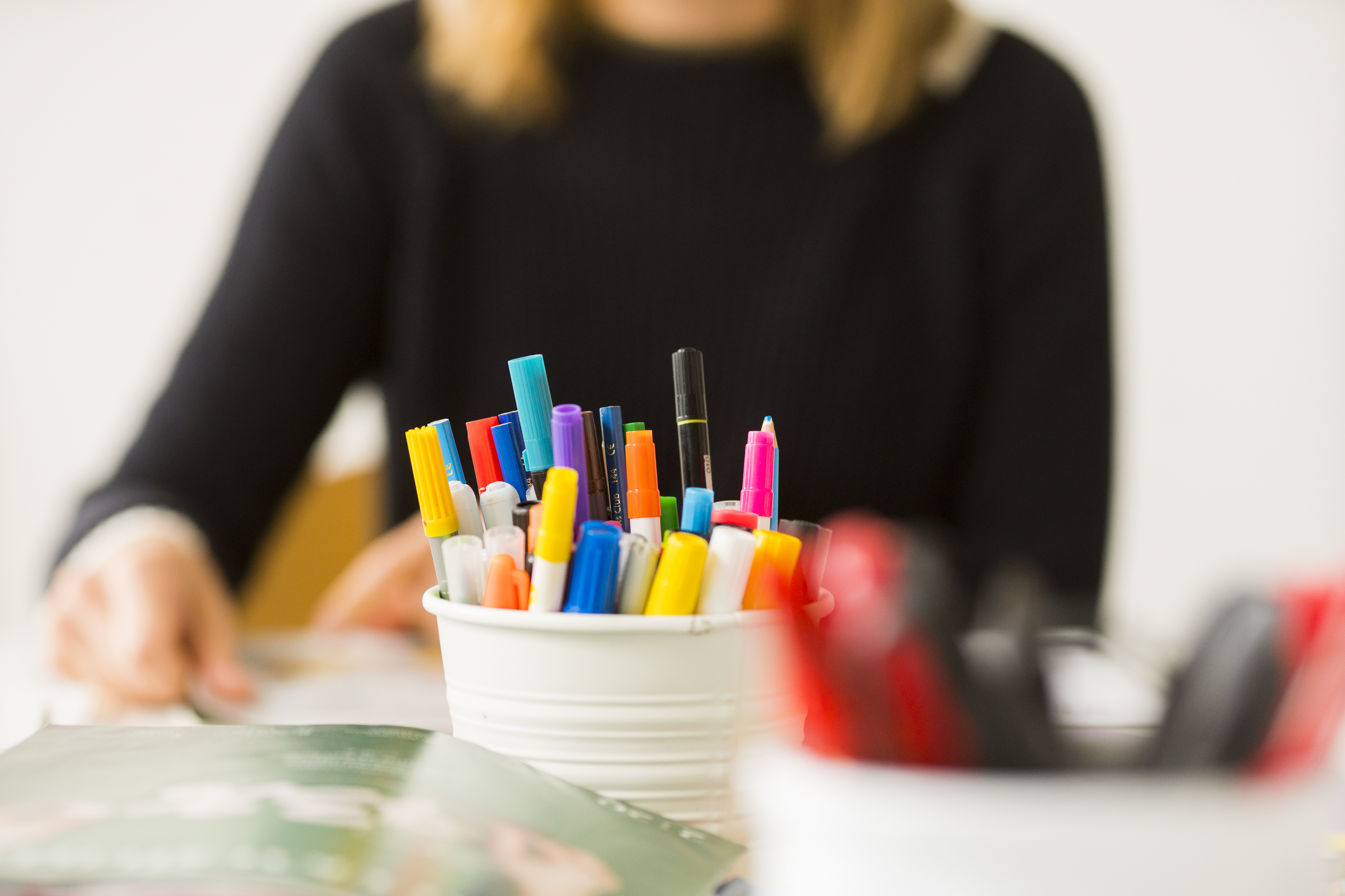 Kuvassa etualalla värikkäitä tusseja purkissa ja taustalla epäselvä henkilö istumassa pöydän ääressä.