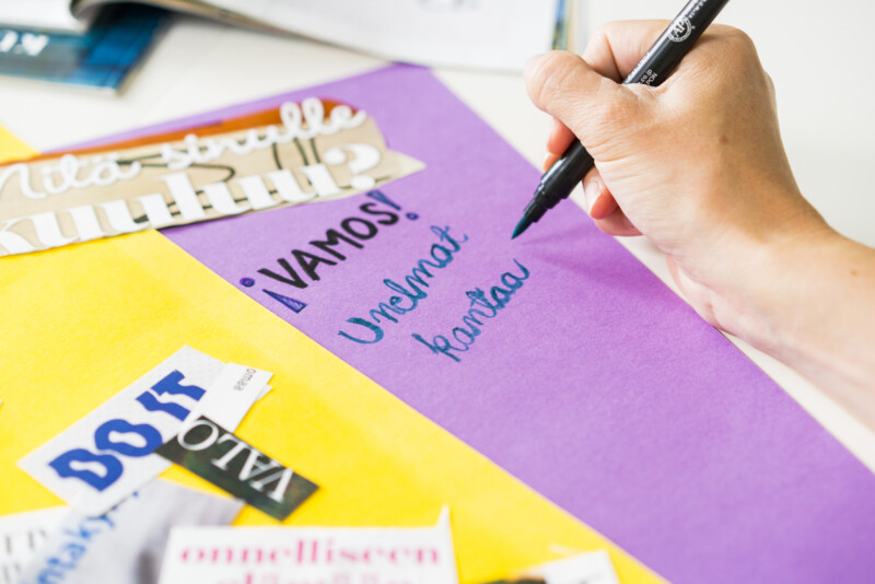 Käsi kirjoittaa mustalla kynällä violettipohjaiseen paperiin tekstiä "VAMOS! Unelmat kantaa" värikkään askartelupöydän ääressä.