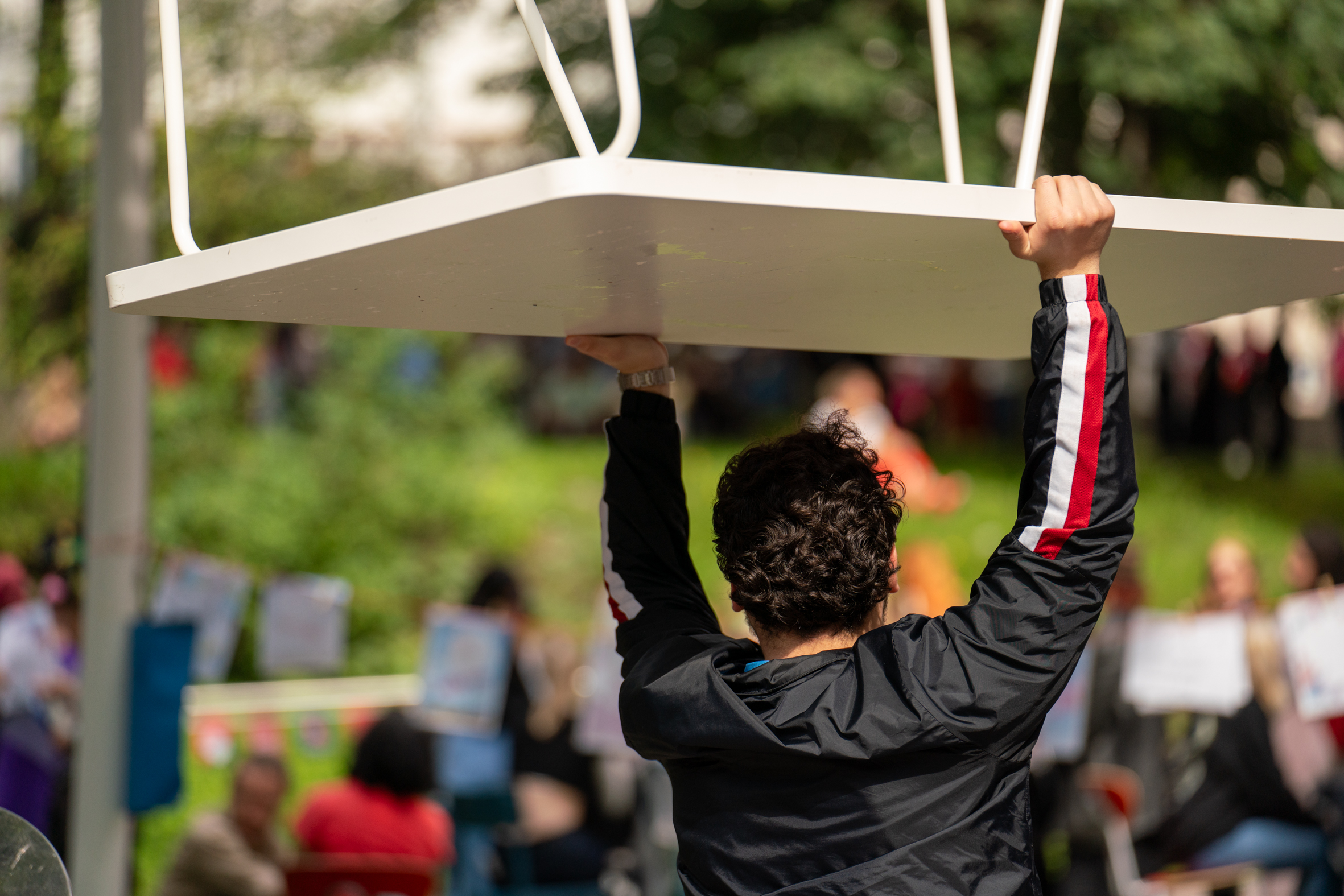 Mies nostaa ylös valkoista pöytää ulkona ihmisten edessä, taustalla vihreää puistoaluetta ja muita ihmisiä.