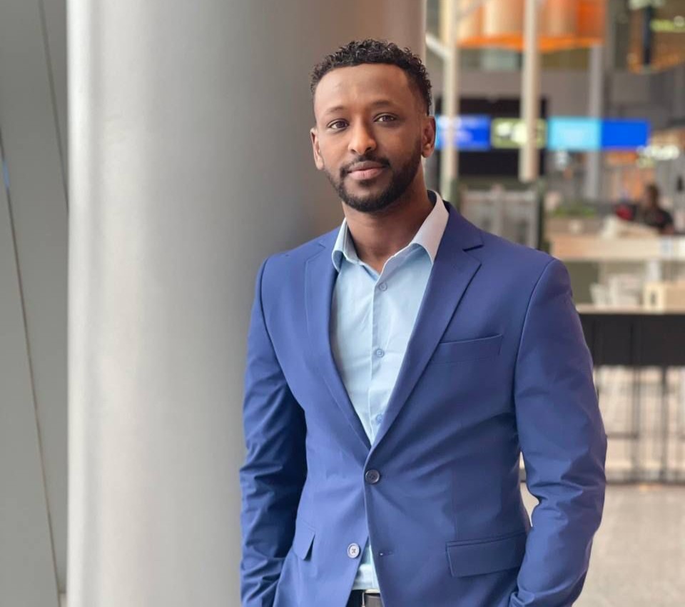 Mies sinisessä puvussa seisoo sisätiloissa, taustalla näkyy moderni julkinen tila ja ihmisiä.