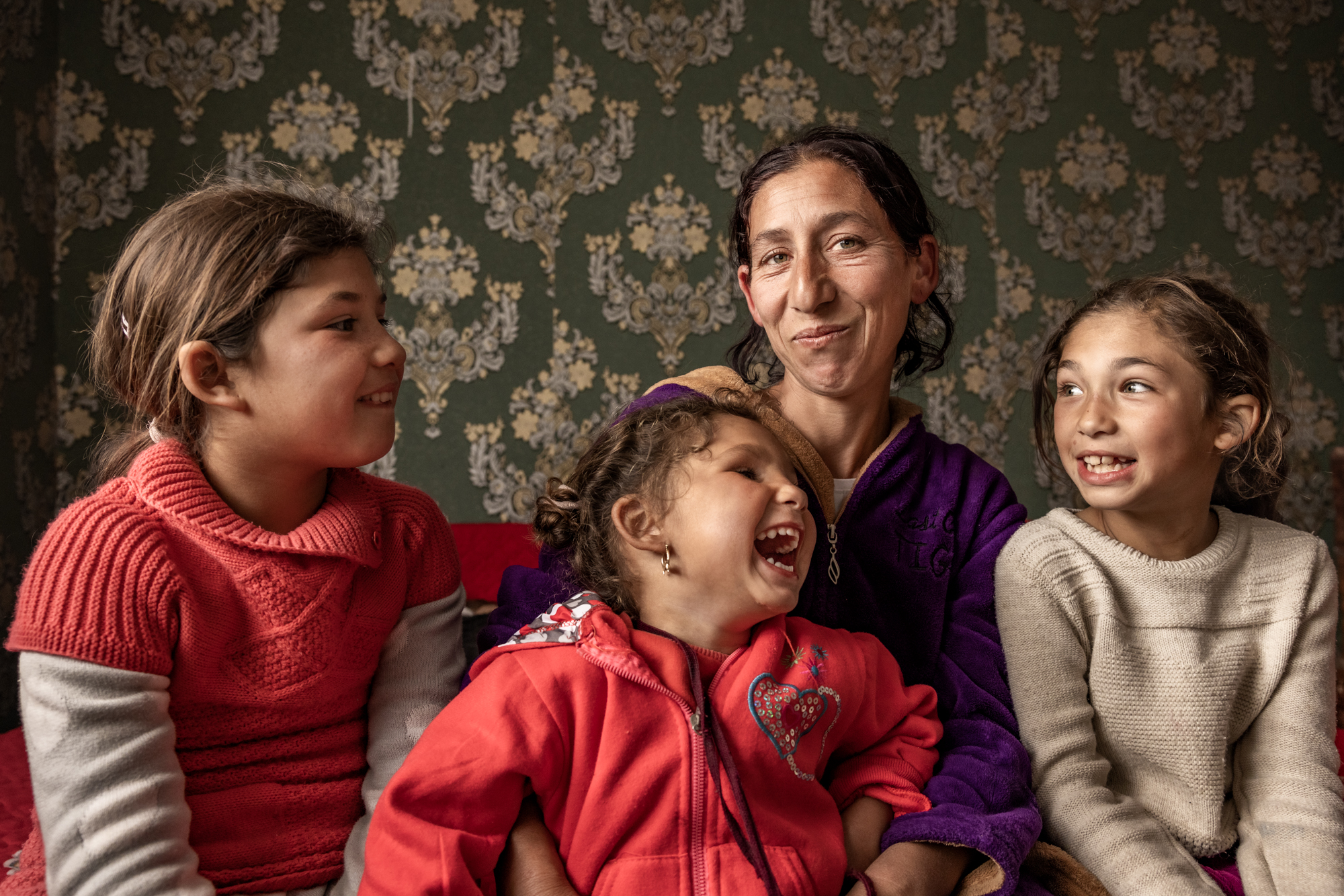 Kolme lasta ja aikuinen istuvat yhdessä hymyillen ja katsovat toisiaan lämpimässä kodinomaisessa ympäristössä.