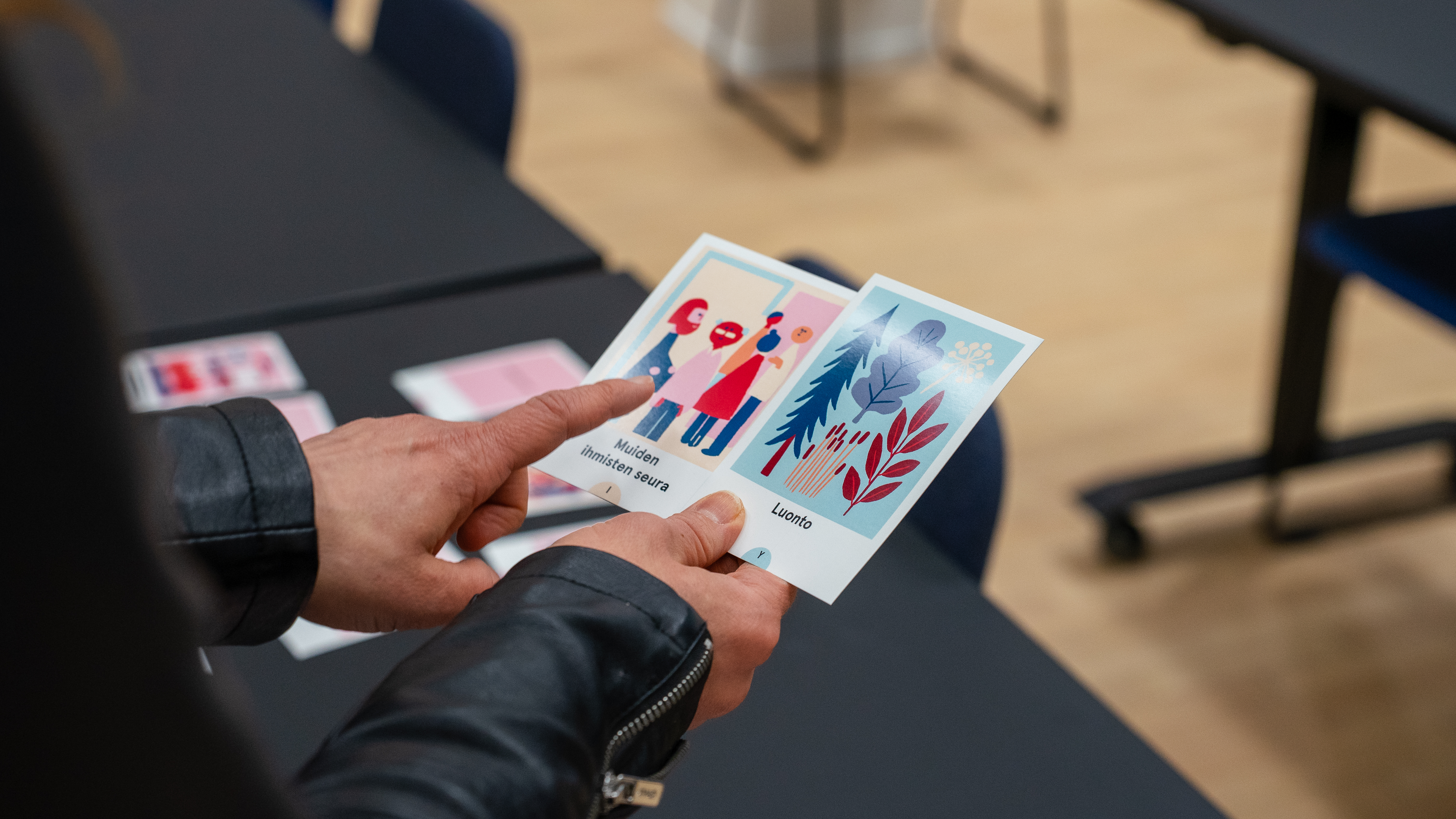 Käsi pitelee ja osoittaa kolmea erilaista korttia, joissa on graafisia kuvituksia, taustalla näkyy pöytä ja tuoleja.